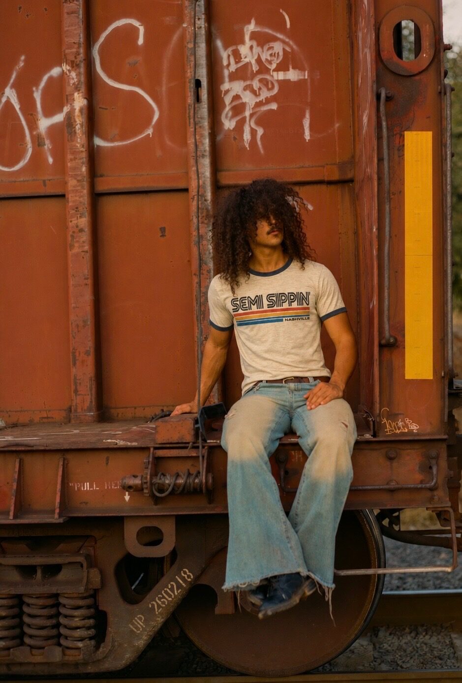 The Nashville ringer tee on a man sitting on the coupling of a rusted freight car