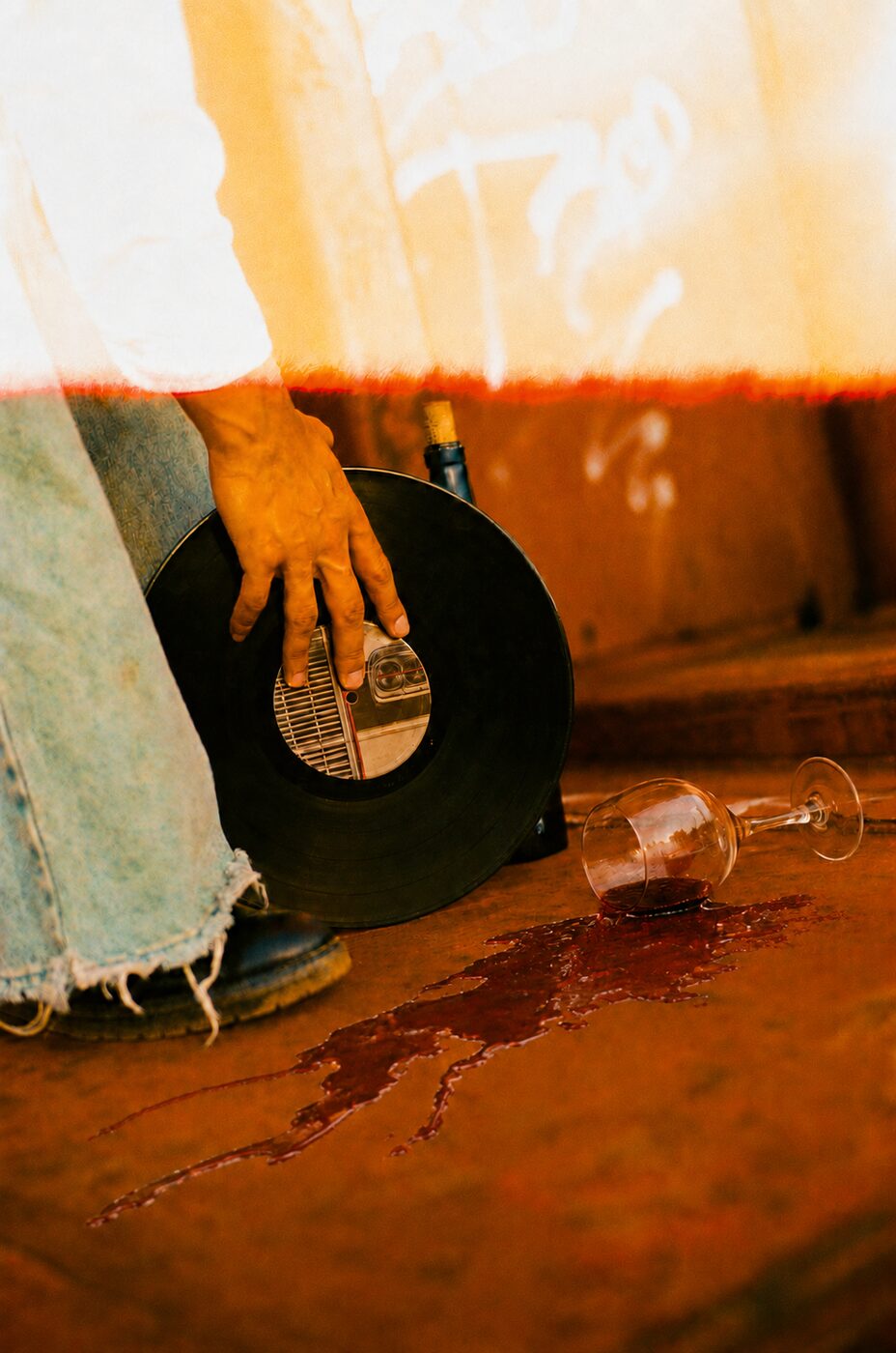 A hand on a vinyl record, a glass of red wine spilled across rust-coloured ground